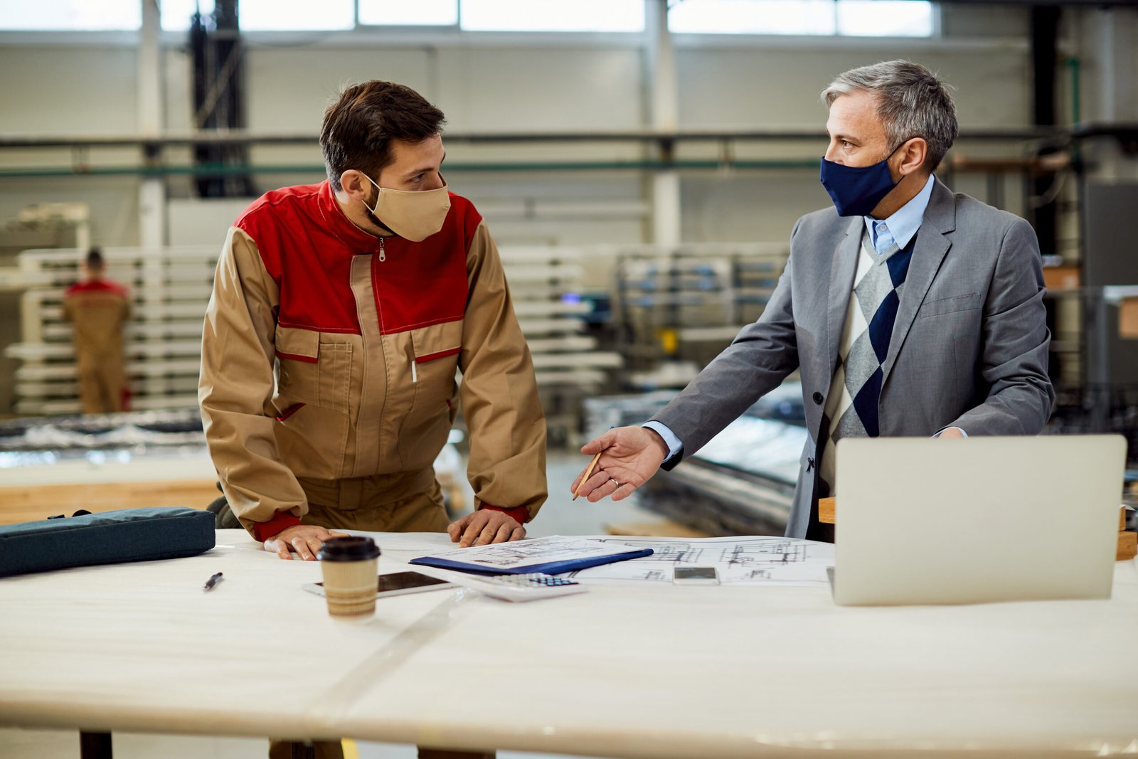Mid adult businessman and male worker discussing about project plans while wearing protective face masks at carpentry workshop.
